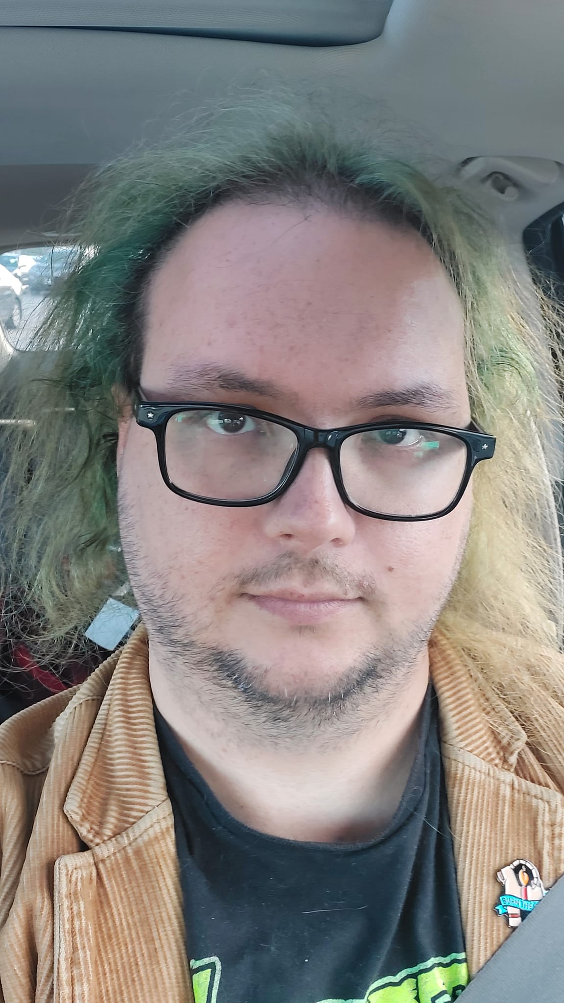 A man with green hair wearing a brown corderoy jacket sits in a car looking slightly uncomfortable